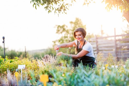 A woman with dark hair tied back, wearing a white shirt and black apron, tending to a colorful garden bed in an outdoor space. She is smiling while using her hands to carefully plant or arrange flowers within a designated flower bed bordered by tidy plants and shrubs. The garden features a variety of greenery, including low-growing plants and flowering bushes, with patches of soil visible and edged by a neat border. In the background, there are taller trees and a wooden fence, suggesting a residential yard in Woolwich. The scene is illuminated by natural sunlight, creating a warm and inviting atmosphere with soft shadows. The setting reflects a well-maintained outdoor area suitable for professional gardening and landscaping services offered by Landscapers Woolwich, highlighting flower bed creation amid a lush, vibrant environment.