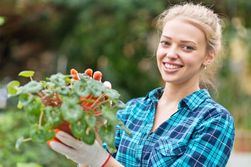 A young woman with blonde hair tied back, smiling while holding a small potted plant with green and reddish leaves in her gardening gloves, standing in a lush backyard garden with a well-maintained lawn, nearby hedge, and flowering plants in the background, under natural daylight in a bright outdoor setting, capturing the natural beauty and care involved in gardening and outdoor landscape maintenance within a typical UK garden environment near Woolwich, highlighting the importance of outdoor plant care and landscape upkeep as part of professional gardening services by Landscapers Woolwich.