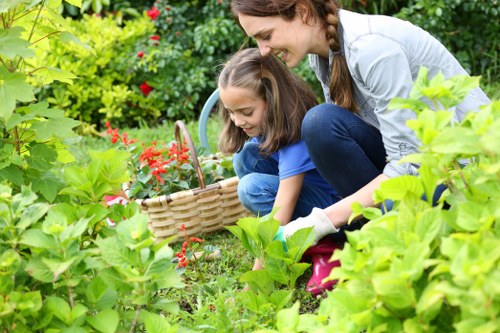 A woman and a young girl are engaged in gardening within a residential garden in Woolwich, surrounded by lush green foliage, flowering plants, and shrubs. The woman is crouched down, smiling, with long hair tied back, wearing a light grey top and jeans. The girl, dressed in a bright blue shirt and jeans, is also crouching and holding a small gardening basket. The garden features a well-maintained lawn with dense grass, bordered by a variety of flowering plants with red blossoms, and edged by leafy green hedges. In the background, there are taller shrubs and trees, providing shade and a natural backdrop, and the overall scene is bathed in soft, natural daylight, suggesting a calm and clear weather. This outdoor space exemplifies typical garden maintenance and planting activities, relevant to professional gardening and landscaping services like those offered by Landscapers Woolwich, in a quiet residential area with a postcode likely in SE London.