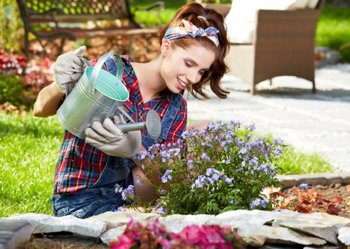 A young woman with brown hair tied back with a colorful headband, wearing a plaid shirt and grey gardening gloves, tending to a flower bed in an outdoor garden setting in Woolwich. She is holding a small metal watering can in her left hand and is smiling as she waters a cluster of delicate purple flowers with green foliage. The flower bed is edged with flat stones, and the surrounding garden features a well-maintained lawn with lush green grass. In the background, there is a paved patio area with outdoor furniture and a wooden fence, suggesting a residential garden space designed for relaxation and outdoor activities. The scene is sunny with natural daylight illuminating the vibrant colors of the flowers, leaves, and the woman's clothing, highlighting the natural textures of soil, plants, and paving surfaces, and reflecting the professional garden maintenance services offered by Landscapers Woolwich in the local area.