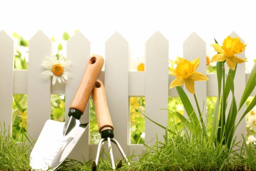 A close-up view of a small garden bordered by a white picket fence, featuring vibrant yellow daffodils and white daisies growing alongside lush green grass and soil. Two gardening tools—a hand trowel and a hand rake—are inserted into the soil in the foreground, suggesting recent planting or maintenance activity. The background shows a blurred garden environment with bright natural sunlight, indicating a clear day in Woolwich. The scene reflects typical outdoor garden setup suitable for professional gardening and planting services offered by Landscapers Woolwich, with attention to plant health, flower display, and tidy layout. The overall composition emphasizes a well-maintained, lively outdoor space within an urban or suburban garden setting, aligning with services related to planting, lawn care, and garden improvement in the local area.