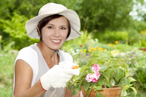 A woman wearing a wide-brimmed white hat, light grey gardening gloves, and a white short-sleeved top is tending to a potted flowering plant in an outdoor garden setting. The garden features a lush, green lawn in the foreground, bordered by various flowering plants and shrubs with pink blooms. In the background, there are tall trees with leafy green canopies, creating a shaded and vibrant environment typical of a well-maintained backyard in Woolwich. The scene is captured on a bright, sunny day, with natural light illuminating the foliage and flowers, highlighting the natural tones of green, pink, and earth within the space. This image exemplifies gardening activities such as planting or caring for flowers, aligning with outdoor garden maintenance services offered by landscapers in Woolwich, focusing on enhancing outdoor aesthetics and plant health.