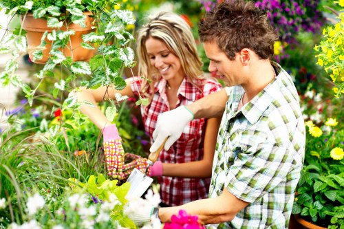 The image shows two individuals working together outdoors in a vibrant garden, with a variety of lush green plants, flowering shrubs, and colorful blooms arranged in flower beds and containers. The garden features a well-maintained lawn area with dense, healthy grass in the foreground, bordered by raised flower beds filled with a mix of flowering plants and decorative foliage. In the background, there are potted plants hanging from a wooden structure, alongside a variety of trees and bushes providing natural shade and structural elements typical of a landscaped outdoor space. Both people are wearing gardening gloves, and they are actively engaged in planting or tending to the plants, with gardening tools visible, highlighting outdoor maintenance activities. The weather appears bright and sunny, illuminating the vivid colours of the flowers and greenery, consistent with outdoor gardening work in Woolwich or nearby areas. The scene demonstrates a cared-for, thoughtfully designed garden environment, reflective of professional gardening and landscaping practices supported by local companies such as Landscapers Woolwich, emphasizing outdoor aesthetic and plants' health.