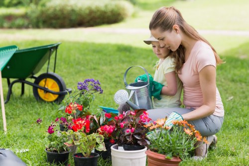 The image shows a woman and a young girl working together in a well-maintained garden, surrounded by a variety of flowering plants and green foliage. They are kneeling on a lush, evenly mowed lawn with vibrant red, pink, purple, and orange flowers planted in pots and flower beds, creating a colourful contrast against the green grass. In the background, a garden wheelbarrow with a yellow wheel is positioned on the grass, indicating ongoing garden maintenance or planting activity. The garden features a mix of flower beds, grass areas, and possibly some shrubbery, typical of outdoor spaces in Woolwich or nearby London areas, with natural daylight illuminating the scene under a clear or partly cloudy sky. This scene exemplifies outdoor garden care and planting activities, aligning with professional gardening services that focus on creating and maintaining attractive, thriving outdoor environments in the Woolwich area, supported by local expertise and landscaping knowledge.