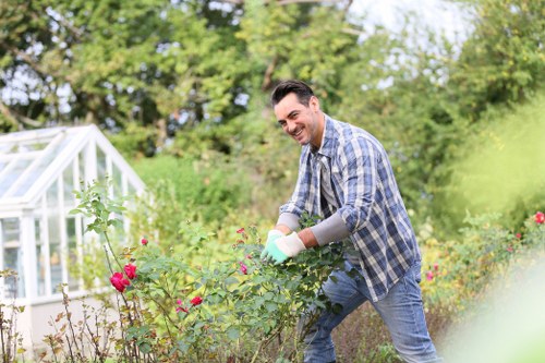 A man is working in a well-maintained garden with lush green trees and shrubs in the background. He is pruning or tending to a flowering shrub with bright pink blossoms, using gardening gloves for protection. The garden features a mix of grassy lawn areas, neatly arranged flower beds, and a paved pathway. To the left, a white greenhouse is partially visible, indicating a space for plant cultivation. The weather appears clear, sunny, and suitable for outdoor gardening activities, contributing to a vibrant and inviting outdoor environment. This scene exemplifies professional outdoor maintenance and gardening services, showcasing care for varied garden elements such as plants and surfaces, consistent with the offerings of Landscapers Woolwich in the Woolwich area, post code SE18, enhancing the natural beauty and health of residential outdoor spaces.
