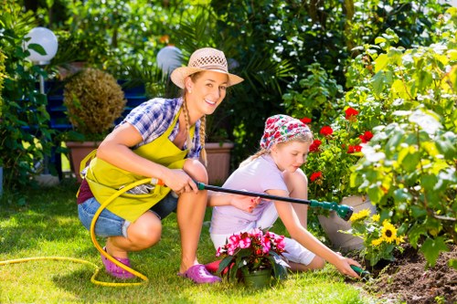 A woman and a young girl are gardening together in a lush backyard garden, surrounded by various plants, flowers, and shrubs. The woman is crouching on the well-maintained grass, wearing a straw hat, a plaid shirt, and a bright yellow apron, holding a garden hose. The girl, dressed in a pink and white patterned hat and light-colored clothing, is planting or tending to flowers in a flower bed bordered by a low stone edge, with vibrant blooms in shades of pink, red, and yellow. In the background, there are bushes, trees, and decorative outdoor lighting, creating a peaceful and inviting outdoor environment suitable for landscaping and garden maintenance. The scene is illuminated by natural sunlight, indicating a clear, pleasant day, emphasizing the outdoor space’s natural beauty and garden care activities conducted by Landscapers Woolwich near the Woolwich area, as part of their gardening services.