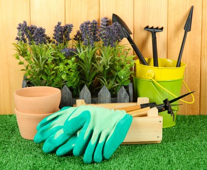 The image features a well-organized outdoor garden scene with various gardening tools and supplies arranged on a patch of artificial green turf. In the background, there are several potted lavender plants with purple flowers and lush green foliage, placed against a wooden fence. To the right, a bright yellow plastic bucket contains long-handled garden tools such as a trowel, spade, and cultivator, all with black handles. In front of the plants, there is a small wooden crate holding a pair of light blue gardening gloves, a small hand rake, and a gardening fork, suggesting readiness for planting or cultivation work. Additionally, a couple of beige terracotta pots are stacked nearby, ready for planting new flowers or shrubs. The scene is well-lit, indicating daylight, and the overall arrangement presents a tidy and functional outdoor gardening setup suitable for maintaining a landscaped garden area in Woolwich or nearby areas, reflecting professional gardening service elements.