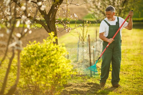 A middle-aged man wearing dark green overalls and a white shirt is standing in a well-maintained garden, holding a garden rake with a blue head and red handle, engaged in outdoor maintenance. The garden features a lush lawn with evenly cut grass and a flower bed bordered by soil, situated near a flowering tree with light pink blossoms. In the background, there is a small wire fence separating different garden sections, with additional trees and shrubs further back, under bright natural sunlight indicating clear weather. The scene reflects typical gardening activities on a sunny spring day in Woolwich, showcasing the outdoor space's landscaped layout including grass, flower beds, and garden tools, consistent with professional gardening services. The environment appears clean, organized, and well cared for, supporting local landscaping and gardening maintenance efforts by Landscapers Woolwich.