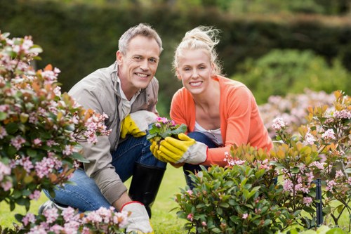 A smiling man and woman are gardening together in a lush outdoor setting, surrounded by flowering shrubs with pink blossoms and dense green foliage in the background. The man has short, styled grey hair and is wearing a light grey shirt and gardening gloves, while the woman has blonde hair tied back and is dressed in an orange top, also wearing gloves. They are crouched down, tending to the plants, with the woman holding garden shears. The garden features a well-maintained lawn with vibrant green grass, and the scene is bathed in natural daylight, indicating a clear, pleasant weather. This outdoor space appears part of a landscaped backyard or front garden, with a focus on flowering plants and shrub borders, characteristic of professional garden maintenance and landscaping projects by companies like Landscapers Woolwich. The setting reflects a healthy, inviting outdoor environment suited for regular gardening services, including planting, pruning, and lawn care, in the Woolwich area postcode SE18.